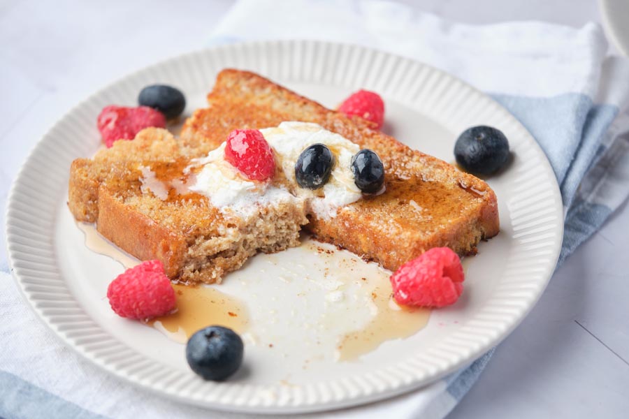 A bite taken out of a slice of keto french toast that is topped with whipped cream, berries and syrup.