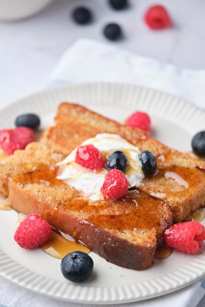 A slice of french toast on a plate with raspberries and blueberries scattered around.