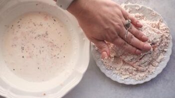 A hand patting a piece of chicken coated with a protein powder breading and a bowl of buttermilk next to it.