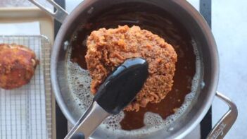 Tongs holding a piece of fried chicken over a pot of hot oil.