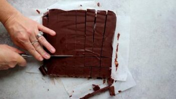 A slab of set fudge being sliced into squares on parchment paper with a knife.