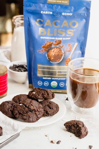 A bag of Cacao Bliss behind a plate of chocolate cookies with a mug of mocha coffee nearby.