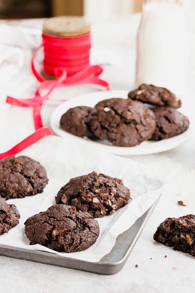 Chocolate cookies sitting on a baking tray with another pile of cookies behind next to red ribbon.