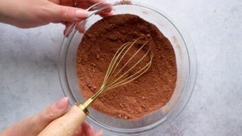 A wire whisk in a clear bowl filled with chocolate dry ingredients.