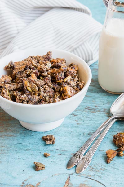 A bowl of granola cereal on a blue table with spoons and milk nearby.