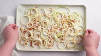 Hands holding a baking sheet with onion rings coated with panko.