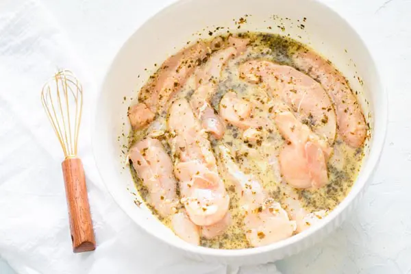 a brown wire whisk next to a white bowl of marinaded chicken