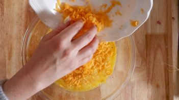 adding shredded cheddar cheese to the egg mixture in a clear bowl