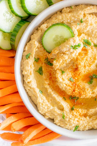 A large bowl of hummus made with lupin beans with a cucumber slice in the dip next to carrot sticks and more cucumber slices.