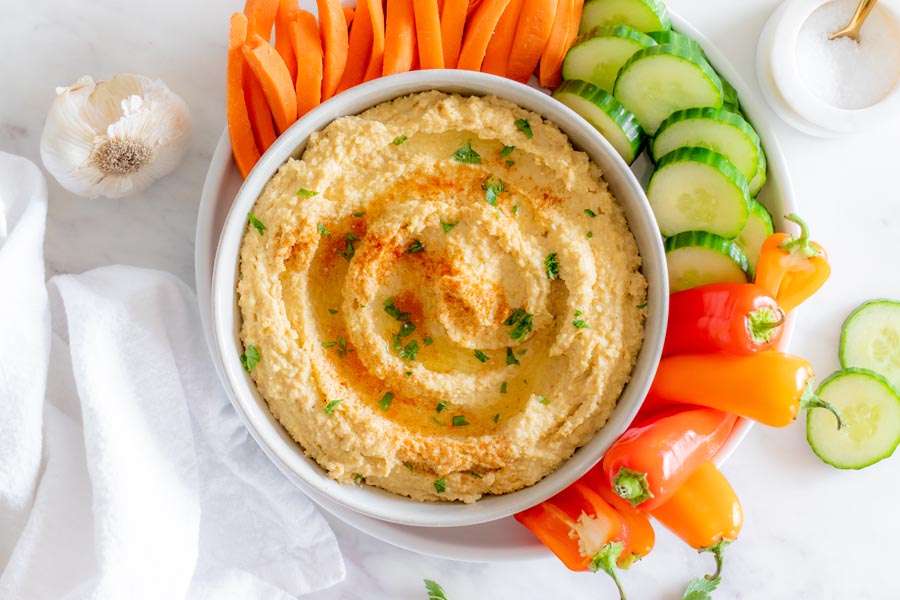 Thick hummus in a white bowl served with sweet peppers, cucumber and carrot and topped with parsley.