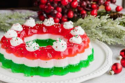 Decorated red, white and green jello salad topped with whipped cream and sprinkles in front of holly berries.