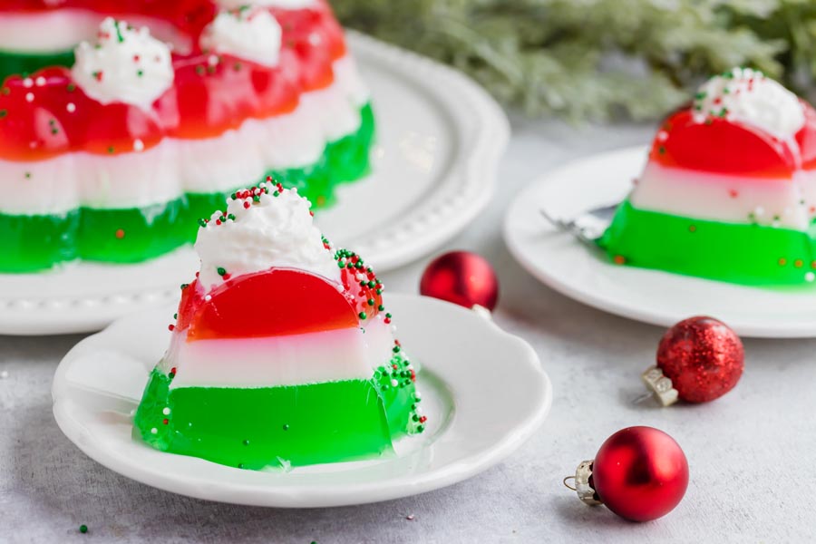 Two slices of Christmas jello salad on small white plates topped with whipped cream and sprinkles with Christmas ornaments nearby.