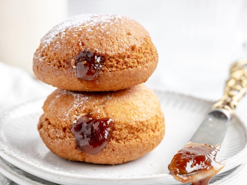 two fried donuts with strawberry jelly inside next to a knife with a smear of jam