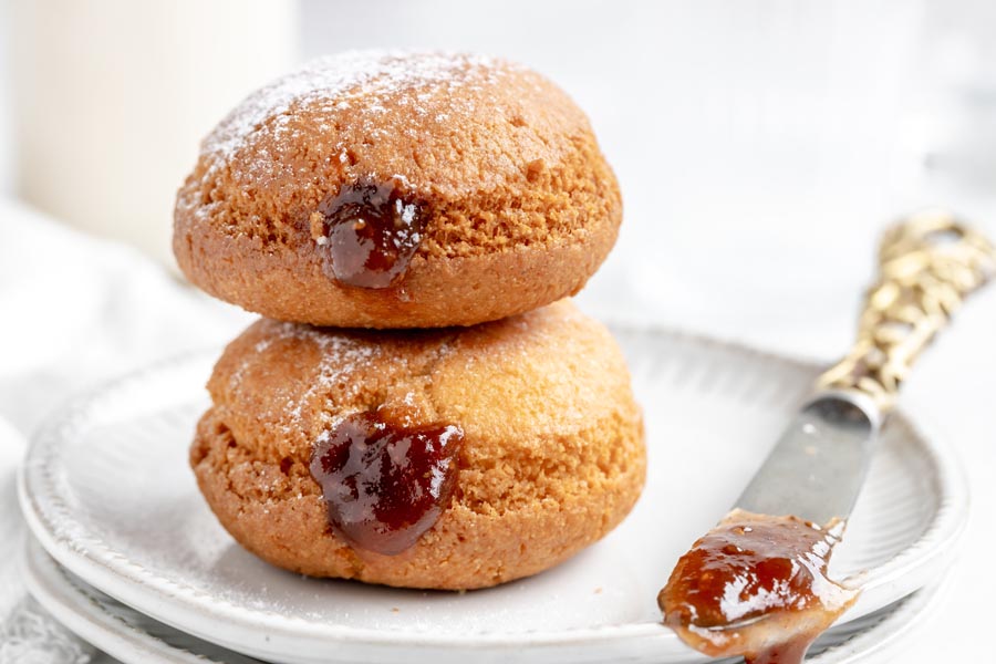 two fried donuts with strawberry jelly inside next to a knife with a smear of jam