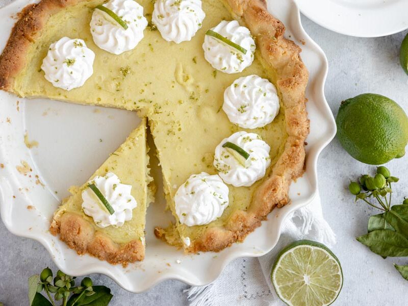 a slice of pie cut out of the whole pie with dollops of whipped cream around and limes tossed around