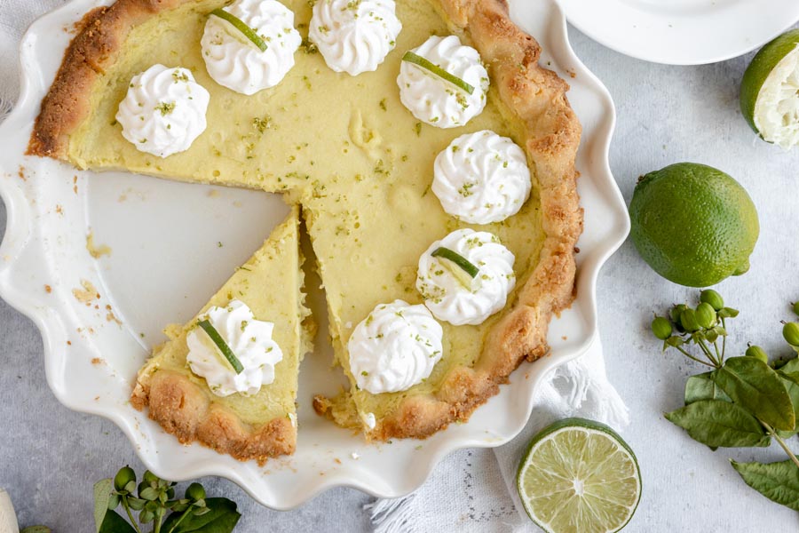 a slice of pie cut out of the whole pie with dollops of whipped cream around and limes tossed around