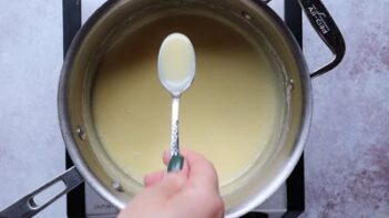 holding a spoon of thicken condensed milk above a sauce pan with more sweetened condensed milk