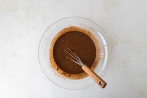 A glass mixing bowl filled with chocolate batter and a wooden-handled whisk resting inside.