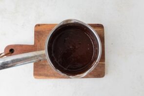 Small saucepan with melted chocolate ganache sitting on a wooden cutting board.