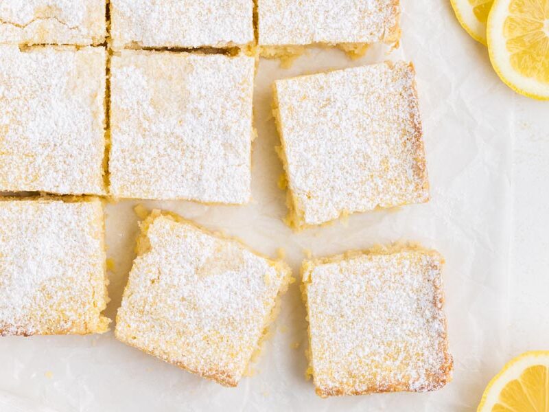 Lemon squares cut up on a board with parchment paper and sliced lemons around.
