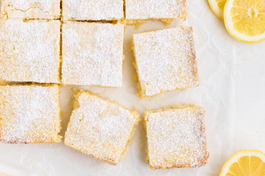 Lemon squares cut up on a board with parchment paper and sliced lemons around.