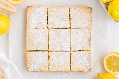 Lemon bars cut into squares on a sheet of parchment paper with lemons around.