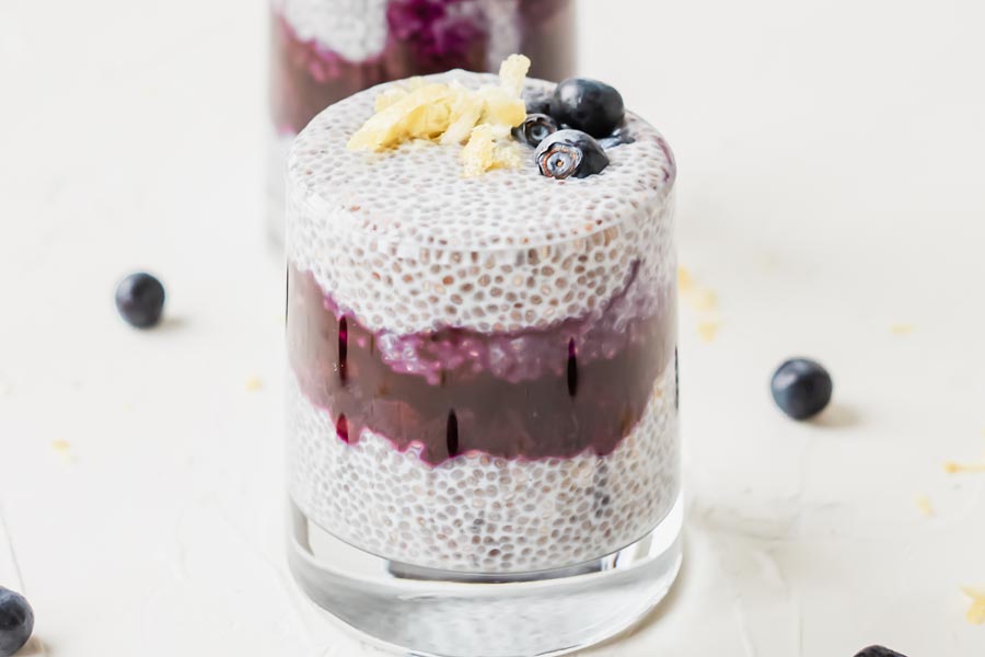 one glass of chia pudding in front of another glass topped with blueberries and a lemon curl