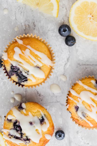 Three glaze topped blueberry muffins next to whole blueberries and lemon slices.