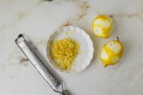 Fresh lemon zest on a small plate beside whole lemons and a microplane grater.