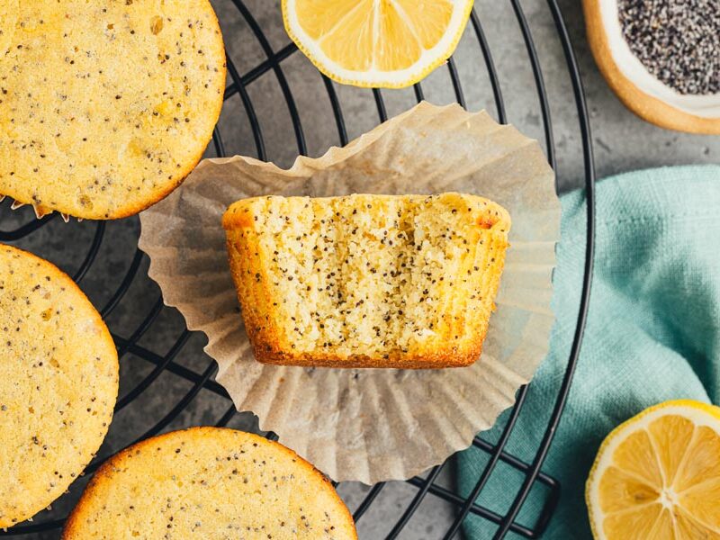 A bite out of lemon poppyseed muffins on a liner next to more muffins on a wire rack.