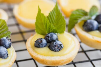 A little bite sized lemon tart encased in a flaky crust and topped with three blueberries and some mint leaves.