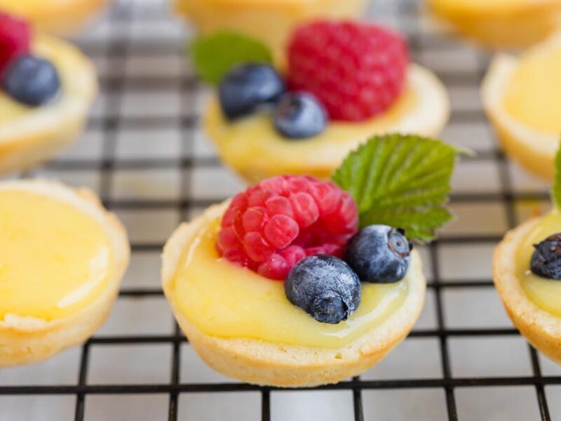 Mini lemon tarts topped with raspberries, blueberries and mint leaves on a cooling rack.