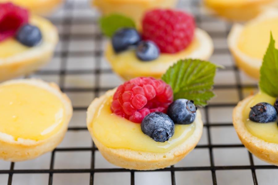 Rows of mini lemon tarts topped with fresh berries - raspberries and blueberries- some have mint leaves.