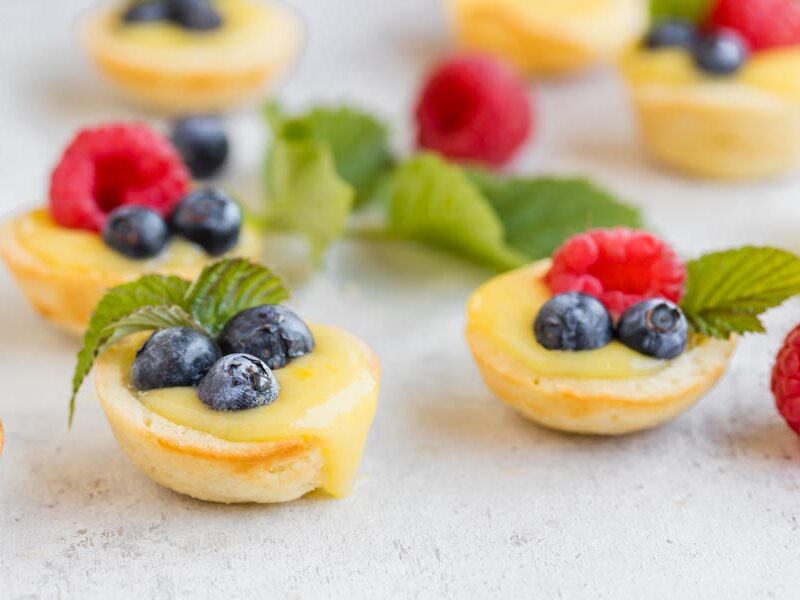 Lemon tarts filled with lemon curd and berries with raspberries and mint leaves scattered around.