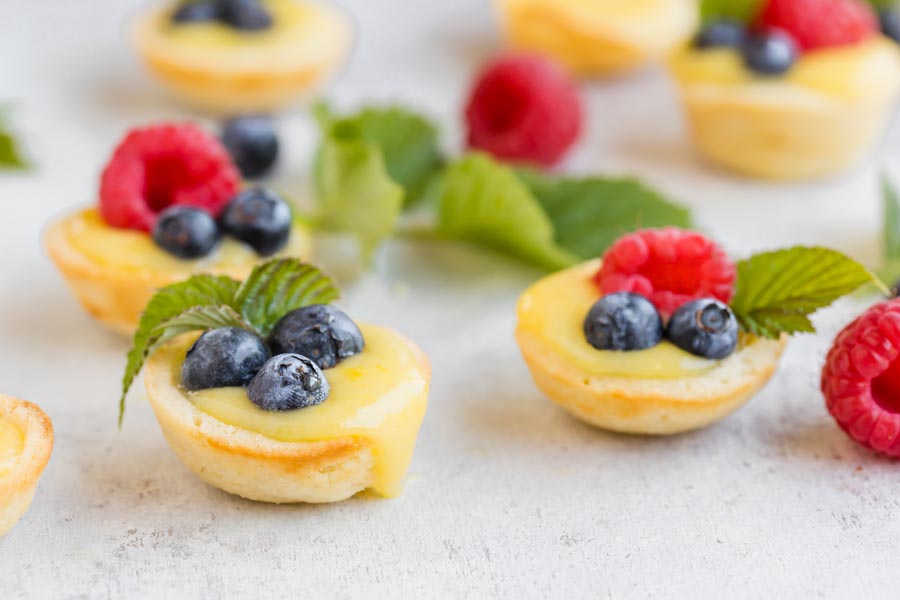 Lemon tarts filled with lemon curd and berries with raspberries and mint leaves scattered around.
