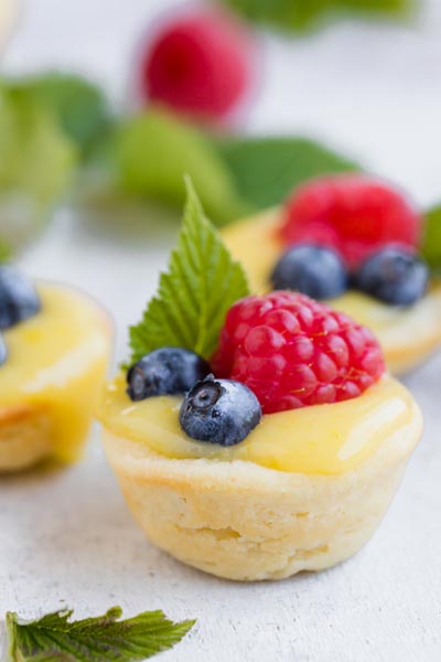 Mini lemon tarts filled with creamy curd and topped with raspberries, blueberries and mint leaves.