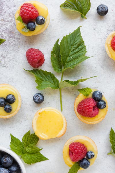 Top down view of bite sized lemon tarts topped with berries with leaves and berries scattered around.
