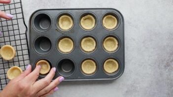 Hands pulling out baked mini pie crusts from a muffin pan and placing them on a wire cooling rack.