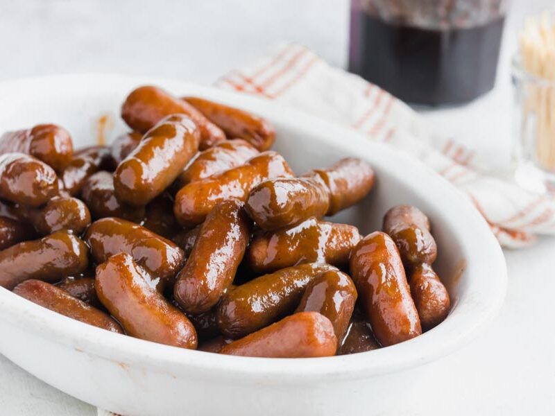 A bowl of cocktail weenies tossed in bbq sauce and grape jelly with toothpicks in the background.
