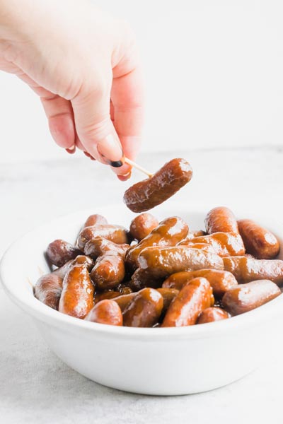 A hand holding a toothpick stabbed into a cocktail sausage.