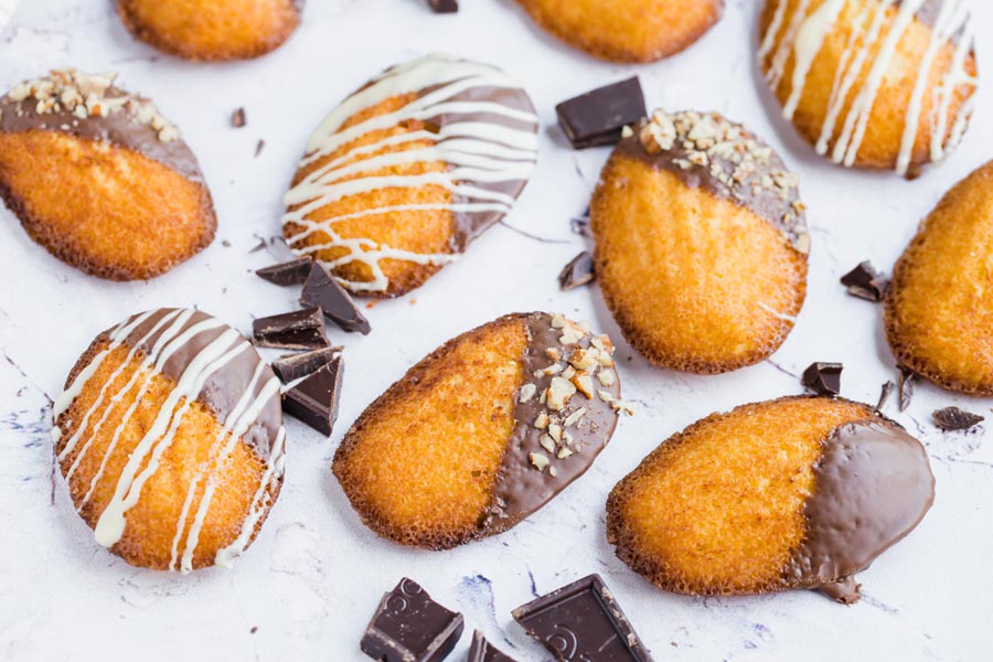 scalloped madeleine cookies covered in chocolate scattered around a table with broken pieces of chocolate