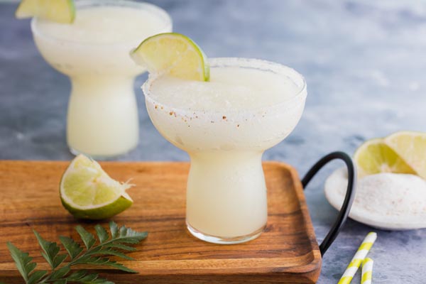 two cocktails one on a wooden plater with a lime and a fern