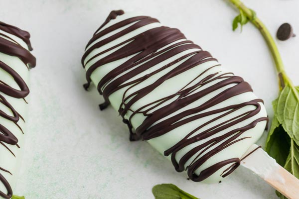 a frozen popsicle coated with chocolate on a counter with mint leaves and chocolate chips