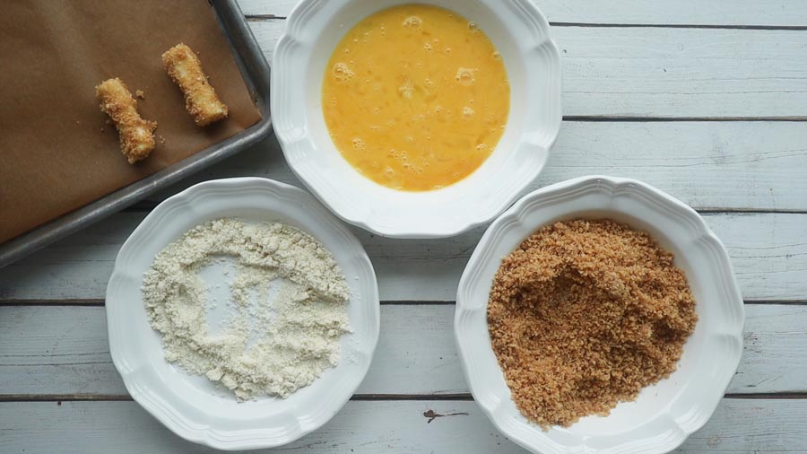 three bowls filled with coating mixtures and two cheese sticks on a baking tray