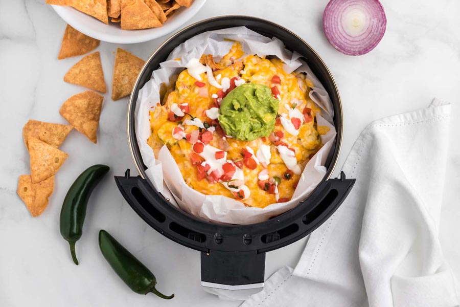 An air fryer basket holding a pile of nachos topped with melted cheese, sour cream and guacamole.