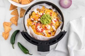 An air fryer basket filled with nachos topped with guac, sour cream, cheese and pico de gallo next to more chips and jalapenos.