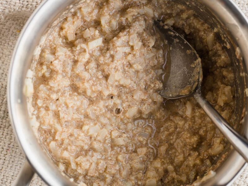 A saucepan with cooked noatmeal inside made from riced cauliflower. A spoon is lying in the pot.