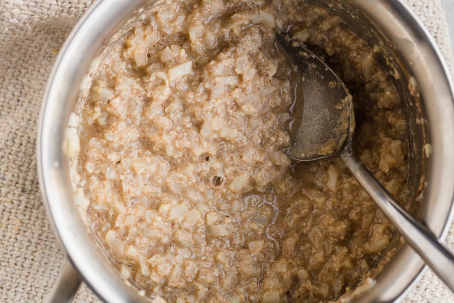 A saucepan with cooked noatmeal inside made from riced cauliflower. A spoon is lying in the pot.