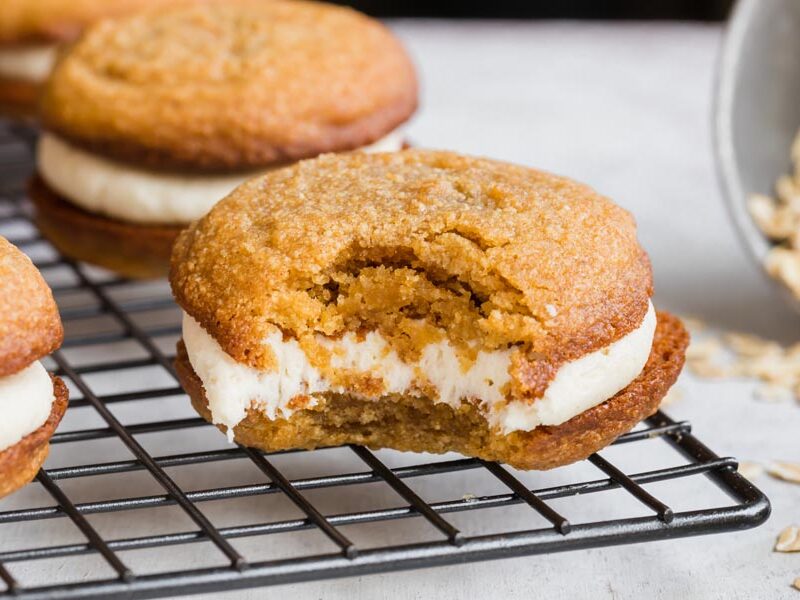 A bite out of an oatmeal cream pie sitting on a wire rack with rolled oats spilled nearby.