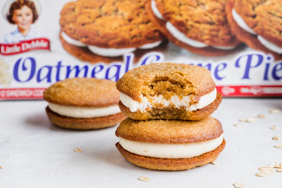 Two homemade oatmeal cream pies stacked on each other with a bite taken out of the top sandwich cookie. A box of Little Debbie's Oatmeal Creme Pies is in the background.